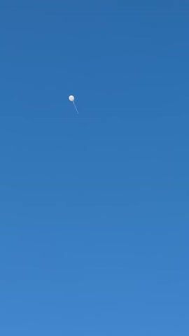 A white, spherical balloon ascends slowly across a clear, bright blue sky in Virši, Latvia. A thin, wavy tail or string hangs from the bottom of the balloon. The sky is free of clouds, indicating fair weather and daytime. The balloon moves from the left side of the frame towards the center as it rises. The only visible subject is the balloon against the vast expanse of the sky. Faint, distant sounds of children's voices can be heard in the background, but no people are visible.