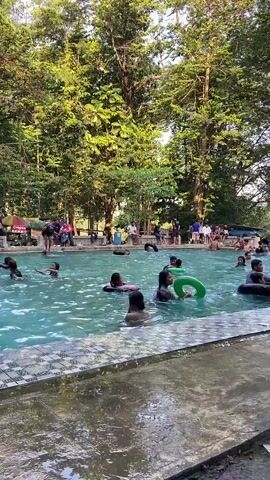 This is a full shot of a swimming pool filled with people of various ages. A group of people are seated on the edge of the pool, and many are in the water, some with inflatable rings. The pool is surrounded by trees, and it appears to be a sunny day. The water is a light blue color and seems to be clean. There are some umbrellas and tables visible in the background, suggesting that this is a public pool or a resort. The overall impression is one of a lively and enjoyable summer day.