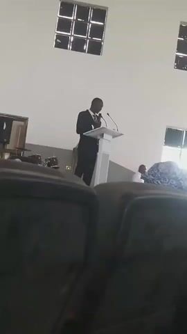 A man in a suit and tie stands at a white podium addressing an unseen audience. The room appears to be a church or auditorium, with chairs visible in the foreground and windows along the back wall. The man is holding a microphone and appears to be speaking or presenting.