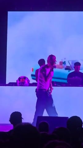 A man wearing a white t-shirt, dark pants, and a red baseball cap is standing on a stage holding a microphone and singing into it. He is gesturing with his hands. Behind him, a large screen displays a blurry image of a blue car and clouds. To his left and right, on a raised platform, are two individuals behind DJ equipment. The person on the left is wearing a purple shirt and looking down at equipment, while the person on the right, wearing a dark shirt, is also looking at equipment. The stage is lit with purple and blue lights, and the audience's silhouettes are visible in the foreground. The overall scene suggests a live music performance.