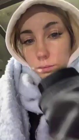 A woman with fair skin and blonde hair is wearing a white hooded sweatshirt and a light blue scarf. She is looking directly at the camera with a slightly downcast expression. The camera angle is from her perspective, and she seems to be holding the camera herself. In the background, there is a soccer field at night. Several people are playing soccer, and there are floodlights illuminating the field. There also appear to be a few tents or structures along the side of the field. The woman seems to be watching the game.