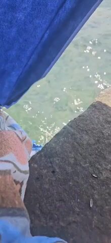 A close-up shot shows a person's legs in blue fabric and a pink and white patterned cloth, leaning over a body of water. The water is clear and shallow, with sunlight reflecting off the surface. A textured, gray rock ledge is visible in the foreground.