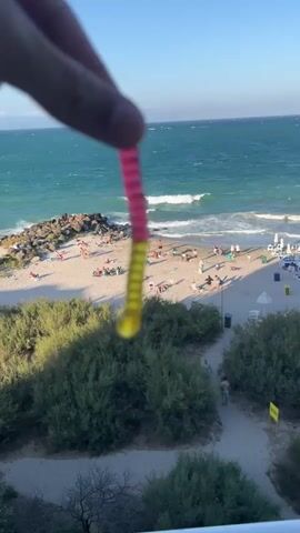 A close-up shot focuses on a pink and yellow gummy worm held between two fingers. In the blurred background, a sunny beach scene unfolds. People are scattered across the sand, some sunbathing, others walking. The turquoise ocean stretches to the horizon, with gentle waves lapping the shore. Lush green bushes and trees line the edge of the beach, leading to a sandy path. The sky is a clear, vibrant blue.