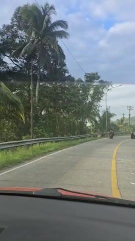 The view from inside a car moving along a two-lane asphalt road shows a landscape with trees and vegetation to the left and right. The sky is partly cloudy. A yellow line divides the road. A motorcycle and several cars are moving in the same direction as the camera. Streetlights and utility poles line the road. In the distance are rolling hills. The dashboard of the car is visible in the foreground.