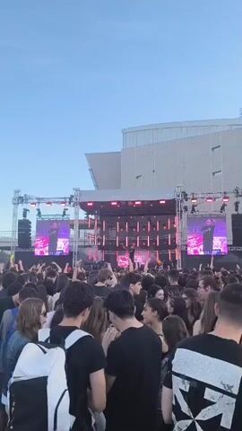 The video shows a dense crowd of people at an outdoor concert. A stage is set up with lighting and large screens displaying an image of the performer. The sky is clear and blue. A large building is visible in the background. Most of the crowd faces the stage. Some people are recording the performance with their phones. The overall atmosphere appears lively and energetic.