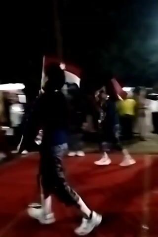 The video shows a nighttime outdoor scene with a group of people gathered around a raised red surface. A large white and red flag is visible in the background, attached to a pole. Several individuals are wearing ornate costumes, some with gold headdresses. One person in the foreground is wearing a dark shirt and blue pants and appears to be taking a photo or video with a device held up to their face. Another person in the background, in a red and gold outfit, is holding a staff or pole. The overall atmosphere suggests a celebration or performance.