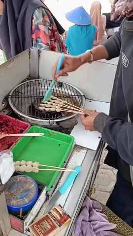 A person wearing a black hooded sweatshirt and a bracelet is grilling meatballs on skewers over a charcoal grill. They are using a turquoise brush to apply a sauce to the meatballs. In front of the grill are containers of red sauce and green sauce, as well as a container of pre-skewered meatballs. To the right of the grill is a pack of cigarettes. In the background, several people wearing hijabs are visible. The scene appears to be outdoors at a street food stall.