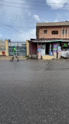 The camera, positioned low and likely mounted on a vehicle, captures a street scene in what appears to be a commercial district on an overcast day. The street is wet, reflecting the diffused light. In the initial shot, a man walks past a row of small shops selling various goods, including school supplies and luggage. A two-story peach-colored building stands behind the shops. As the camera moves, a red van passes, followed by a yellow and black mini-bus. Overhead power lines are visible. The background includes other buildings, shops, and a gated area. The video appears to be a short, unedited clip capturing a typical day in this location.