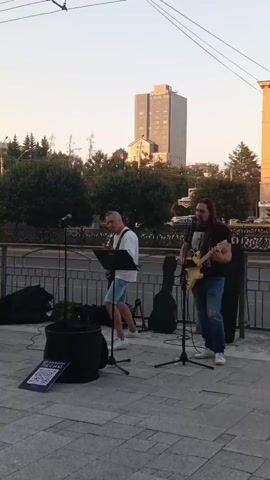 Two men are performing music on a sidewalk. The man on the left is singing into a microphone and holding a sheet of music on a stand. He has gray hair and is wearing a white t-shirt and blue shorts. The man on the right is playing a yellow electric guitar and singing into a microphone. He has long brown hair and is wearing a black t-shirt and ripped blue jeans. Two guitar cases are leaning against a railing behind them. A large black bag is on the ground to the left, and a dark purple bin with a QR code sticker on it is in front of it. In the background, there are trees, a tall building, and traffic. The sun appears to be setting, casting a warm light on the scene.
