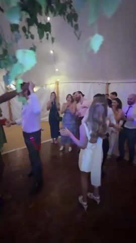 The video shows a group of people dancing at an indoor event, possibly a wedding reception, under a tent. The lighting changes from white to pink and purple. In the foreground, a couple dances closely, with the woman wearing a short, cream-colored dress and the man in a white shirt and black pants. In the background, other guests are visible, some dancing, some watching, and some holding drinks. There are string lights hanging from the ceiling and greenery at the top left of the frame. The floor is made of wood. Another couple is seen dancing in the middle ground with the woman in a black sequined dress and the man in a white shirt and dark pants with red suspenders.