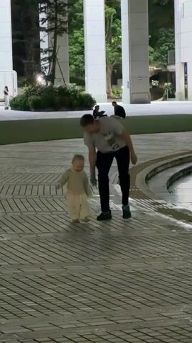 A person in a grey t-shirt and dark pants walks to the right, holding the hand of a small child wearing a light-colored outfit. The child is walking to the right, occasionally looking back at the person. They are on a paved surface with a pattern of diagonal lines. In the background, there are large white columns, trees, and other people sitting or standing. The area appears to be an outdoor public space, possibly at dusk or in the evening, as indicated by the lighting.