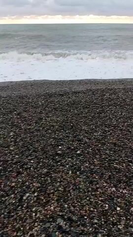 The video shows a pebbled beach on an overcast day. The beach is covered in small, dark pebbles, and the waves from the sea reach the shore. The water is a grayish-green, and the sky is filled with gray clouds, with a small sliver of light visible near the horizon. There are no people or other objects visible in the video, the focus is on the natural scene of the beach and sea.