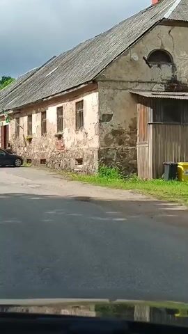 The video shows a drive-by view of a village street, with old buildings and parked cars. The camera pans from a long, old building with a worn facade and a green flag, to a wider shot of the street with other cars parked. Trees and a dark sky are visible in the background. An "ESAM VALA OPEN" sign is also visible on the side of the road.