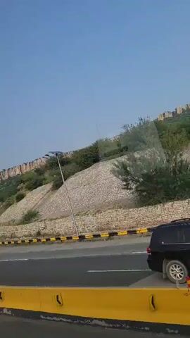 The video shows a convoy of buses on a highway. The buses are white, red, and green, and they appear to be traveling in the same direction. There is also a police car at the front of the convoy. The highway is flanked by a steep, rocky hillside with vegetation. The sky is clear and blue. The video is shot from inside another vehicle, possibly a bus, that is traveling alongside the convoy.