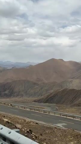 The video shows a winding road through a mountainous landscape under a cloudy sky. The mountains are various shades of brown and grey. The road is asphalt with yellow lines and a grey metal guardrail. In the distance, some snow-capped mountains can be seen. Several vehicles are visible on the road.
