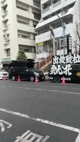 The video shows a street scene in Japan. Several vehicles are parked along the side of the road, including white and black vans, as well as a silver Alfa Romeo. Orange traffic cones are placed around the vans. Several apartment buildings are visible in the background. There is a blue truck and a person walking behind it. Street signs and trees are also visible.