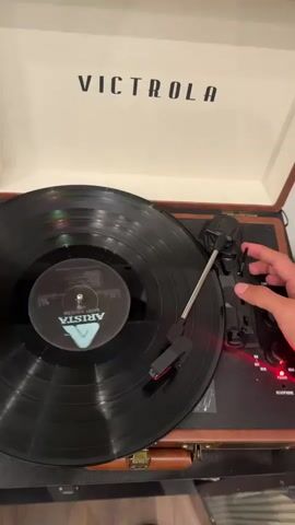 The video shows a close-up shot of a Victrola record player. A vinyl record is on the turntable and the needle is on the record. A hand is seen pressing buttons on the side of the record player. The Victrola logo is visible on the lid of the record player. The record is black with a label that says "Arista." The lighting suggests an indoor environment.