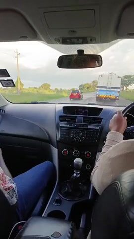 A person driving a car overtakes a red car on a road. The road has trees to the side. The car in which the video is recorded has a manual transmission. There is a passenger in the car wearing blue jeans. A blue and white truck is in front of the driver on the road. The video is recorded from the driver's point of view. The sky is cloudy.