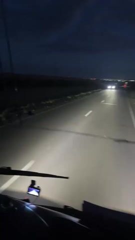 This is a first-person perspective video clip of driving on a road at night. The primary subject is the road itself, illuminated by the vehicle's headlights. The setting is a rural or semi-rural area, with dark fields and sparse vegetation visible on either side of the road. In the distance, a faint line of lights suggests a town or city. The sky is dark with some clouds, indicating it is nighttime. The weather appears clear despite the clouds. There are no visible people in the frame, but there is a dashcam mounted on the dashboard. The activity is the act of driving. Notable details include the bright headlights cutting through the darkness, the white lines on the road, and the distant lights. The overall mood is one of solitary travel through the night. There is no visible text in the video.