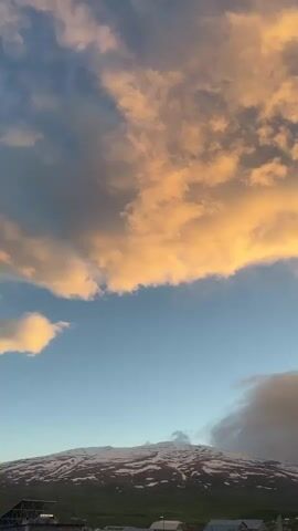 The video shows a scenic view of a mountainous landscape under a partly cloudy sky. The sky is divided into three distinct layers: the top layer consists of golden-orange clouds, the middle layer is a clear blue sky, and the bottom layer contains darker, grayish clouds. A mountain range with patches of snow is visible in the distance, with green fields and some buildings at the base of the mountain. The wind appears to be strong, as indicated by the sound.