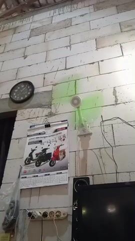 The video opens with a shot of an interior wall made of white, unplastered bricks. A black analog clock, showing approximately 11:55, is mounted on the wall to the left. A small white clip-on fan, emitting a green light as its blades spin, is attached to the wall. Below the fan, a calendar featuring images of motorcycles and scooters hangs. The calendar has text that reads "HEMAT BBM DENGAN SEPEDA LISTRIK" (Save fuel with electric bicycle) and "GENTA." A power strip with multiple outlets is visible near the bottom of the wall, and a black speaker or electronic device is placed on the floor beneath it.

The camera then pans slightly to the left, revealing a doorway or opening covered by dark brown curtains. A young girl, with dark hair tied back, wearing a white tank top and patterned shorts, stands between the curtain panels. She appears to be interacting with or adjusting the gold tassel used to tie back one of the curtains. The room is brightly lit, indicating it is daytime. The exposed brick walls suggest a simple or unfinished interior setting. The location is Baron, Indonesia.
