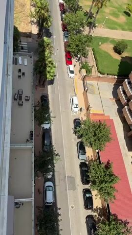 This is an aerial, high-angle, vertical shot looking down on a street lined with parked cars and trees. On the left side of the street, there are buildings, with balconies visible. On the right side of the street, there is a park with grass and palm trees, and more buildings are visible in the background. The sky is clear and the sun is shining. The video pans up and to the right, revealing a wider view of the neighborhood with more buildings and palm trees.