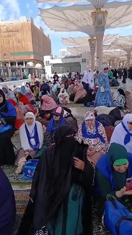 The video shows a crowd of women, mostly elderly, sitting on prayer rugs outdoors, under large white umbrella structures. Many are wearing headscarves and some are wearing niqabs. Some people are looking at their phones, while others seem to be resting or observing the surroundings. There is a building in the background with the words "AL SALAM MUSEUM" inscribed on it. Other structures and people are visible further in the background. The sky is partly cloudy, and the overall scene appears to be a public gathering in an outdoor area.