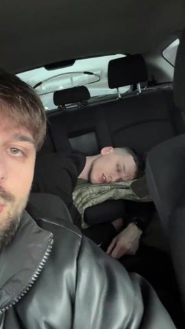 The video opens with a close-up selfie shot from inside a car, presumably being taken by the man in the front passenger seat. He has short brown hair with bangs, a beard, and is wearing a black puffer jacket with a zipper partially open. His right eye is visible, looking directly at the camera.

In the background, visible through the gap between the front seats, another man is sleeping in the back seat. He is lying on his left side, with his head resting on a dark-colored blanket or pillow. He has short dark hair and appears to be wearing a black long-sleeved shirt. His mouth is slightly open as he sleeps. The exterior view through the car's rear window shows a somewhat blurry, grey sky, suggesting the car is in motion.

At the 0:02 mark, the camera pans slightly to the right, revealing a second man sitting in the driver's seat. He has short dark hair, a beard, and is wearing a black hoodie with a white logo on the chest (partially obscured, but appears to be a stylized "NC" or similar). He is wearing a seatbelt and is looking towards the camera with a slight smile, his mouth open as if speaking or reacting to something. The man filming continues to look at the camera, his expression neutral. The sleeping man is still visible in the back, unaffected. The car interior is dark-colored, and the headrests of the front seats are visible.