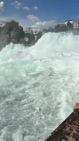 This media file captures a dynamic scene of the Rhine Falls in Dachsen, Switzerland. The primary subject is the powerful waterfall itself, with its churning, white-foamed water dominating the foreground and mid-ground. The water is a vibrant turquoise-green where it's less agitated, contrasting with the frothy white of the cascades and rapids.

In the background, several notable elements are visible. To the left, a rocky outcrop rises, topped with lush greenery and some construction equipment, including a yellow crane, suggesting ongoing development or maintenance. Further back, buildings, including modern high-rise structures and smaller houses, are nestled among trees on a hillside. A Swiss flag is prominently displayed on a rocky peak to the right, adding a patriotic touch to the landscape.

The activity depicted is the raw, energetic flow of the waterfall. The sheer volume of water and the intense turbulence convey a sense of natural power and spectacle.

The time of day appears to be daytime, with bright sunlight illuminating the scene, casting clear shadows and highlighting the textures of the water and rocks. The sky is a mix of blue and scattered white clouds, indicating fair weather. The presence of the Swiss flag and the distinctive architecture in the background strongly suggest the location is indeed Dachsen, Switzerland, home to the Rhine Falls. While there are no visible people clearly identifiable in the frame, the existence of buildings and construction suggests human presence and interaction with the environment. The overall emotion evoked is one of awe at the natural beauty and power of the waterfall. No legible text is visible in the media file.