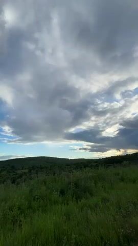 The video shows a panoramic view of a rural landscape with rolling hills covered in green vegetation. The sky is filled with dramatic clouds, with patches of blue sky visible in between. A road winds through the landscape in the foreground, and some distant trees and structures can be seen in the background. The lighting suggests it is either dawn or dusk, with a soft, diffused light illuminating the scene.