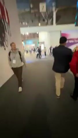 The video shows a busy convention hall with many people walking around. There is a large LED screen that displays abstract patterns and shapes with blue, purple, and white colors. People are walking past the screen, some with luggage. The hall is brightly lit and there are various booths and displays in the background. The floor is dark gray and the walls are white.
