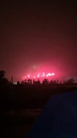 A fireworks display is visible in the distance, illuminating the cloudy night sky with pink and white bursts. The foreground is dark, with the top corner of a blue object, possibly a tent or umbrella, visible. Trees and a tall structure, possibly a smokestack, are silhouetted against the distant lights. The overall scene is dark and atmospheric, suggesting an outdoor event taking place at night.
