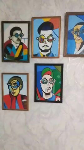 The video shows a wall adorned with five framed portraits. The portraits are abstract and stylized, each depicting a man's face with exaggerated features and bold, geometric patterns in the background. All of the subjects are wearing glasses. The frames are of varying colors and materials, with some being wooden and others appearing to be darker. The wall itself has a textured, light-colored finish. The lighting is consistent, suggesting an indoor environment. There are no people visible in the video.