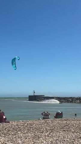 A kiteboarder is in mid-air, having launched from the water. The kite is blue and white and is clearly visible against the bright blue sky. Below the kiteboarder, the sea is a choppy grey-green, with waves crashing against a stone pier. There are several people sitting on a pebble beach in the foreground, some in beach chairs and others lying down. The water appears to be shallow near the shore, with a few people wading in. The weather is sunny and clear, suggesting a warm day at the beach.