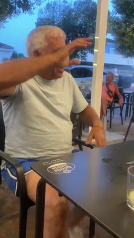 A man with white hair and a light blue t-shirt sits at a black table. He is looking to his left and smiling. The table has a logo on it. There is a glass with a slice of lemon in it on the table. In the background, another man with orange shorts and a black top is sitting at a table. There are trees and buildings in the background. The sky appears to be dusk.