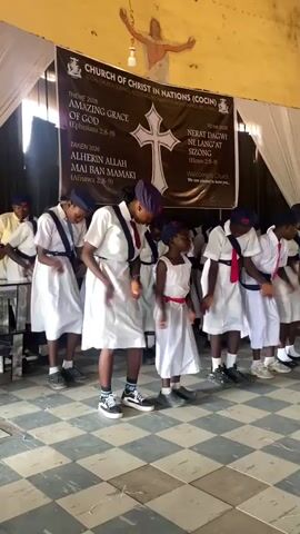 The video shows a group of children in uniform at a church. The children, all girls, wear white dresses with navy blue berets and red neckties. They are lined up in front of a banner for the "Church of Christ in Nations (COCIN)." The banner's theme is "Amazing Grace of God." The children are performing a dance or routine, moving rhythmically to music that can be heard in the background. They hold their hands together in front of them and sway back and forth in unison. Behind them, some adults are seated and appear to be watching the performance. The church has a checkerboard tile floor and a simple, somewhat rustic interior. The atmosphere seems joyful and celebratory.