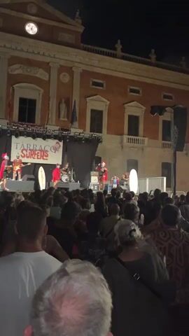 A band performs on a stage at night in front of a crowd of people. The band members are dressed as Roman soldiers, wearing helmets, red capes, and tunics. One member sings and plays a guitar, another plays the drums, and a third is visible in the background playing a guitar. The stage is lit with spotlights and has a large banner behind the band that reads "TARRAGO SURFERS." To the right of the stage, a large white surfboard is standing upright. The crowd is visible in the foreground, mostly in silhouette. The building behind the stage appears to be an old, ornate structure with windows and balconies.