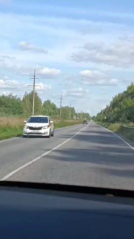 The video shows a view from the interior of a car as it travels down a paved road. The road is bordered by tall grasses and trees on the left side, and more trees and a metal guardrail on the right. Several tall utility poles with wires stretch along the left side of the road. The sky is blue with scattered white clouds. A few other cars are visible on the road ahead and to the right. The interior of the car, including the dashboard, is visible in the foreground. The video captures a sunny daytime scene.