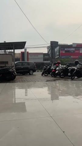 A stationary, eye-level vertical video captures an outdoor scene with a focus on a row of parked motorcycles. The foreground is a wet, tiled surface reflecting the sky and surrounding structures. In the midground, several motorcycles are parked in a line, some with helmets on the seats. A black SUV is parked to the left of the motorcycles. Behind the vehicles, a multi-story building with various signs and advertisements is visible. The sky is overcast and appears bright. The overall atmosphere suggests a cloudy or possibly rainy day.