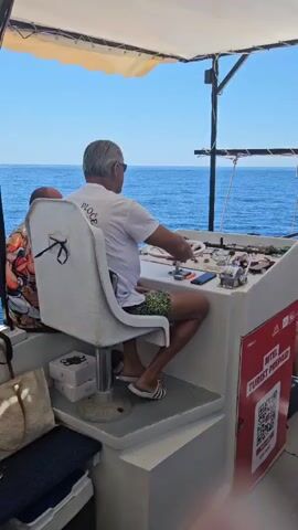 The video shows a man from the back, seated at the helm of a boat. He is wearing a white t-shirt, patterned shorts, and flip-flops. The man has grey hair and is wearing sunglasses. He appears to be steering the boat as he is looking out at the blue ocean. There are various controls and equipment on the console in front of him. To his left, a woman with colorful clothing on is also seated. In the foreground, there are bags and boxes. The scene is set on a bright, sunny day.