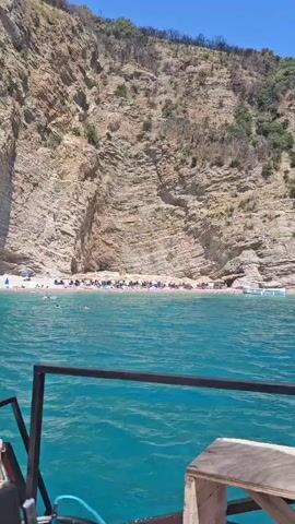 The video shows a view from a boat looking towards a rocky cliff face and a beach. The water is a vibrant turquoise. The cliff is steep and layered, with some vegetation growing at the top. At the base of the cliff is a sandy beach lined with beach umbrellas and chairs. Several people are visible on the beach, sunbathing and walking. In the water, there are more people swimming or wading. A few boats are also present near the shore. The sky is clear and blue. The perspective suggests the boat is moving slowly along the coastline.