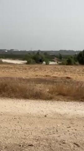 The video shows a drone taking off in a rural setting. The ground is mostly dry, brown grass, with a dirt road in the foreground. In the distance, there are trees, fields, and buildings under a bright sky. The drone ascends into the air, moving away from the camera.