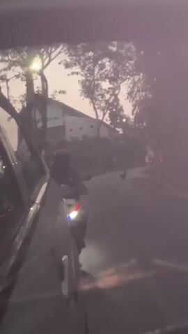 A video taken from inside a car looking out the passenger side window. A person on a motorcycle is visible in the foreground, leaning towards the car. Several other vehicles, including motorcycles and cars, are on the road ahead. The scene appears to be at dusk or early morning, with trees and buildings visible in the background. The road seems to be curving and surrounded by vegetation. There is some haze or fog in the air, and the lighting is dim.