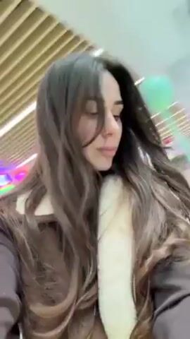 A person with long, dark brown hair, wearing a brown jacket with a white fur collar, is walking and turning towards the camera. The background includes a glass window with blue sky and architectural structures visible through it, suggesting an indoor location such as a mall or airport.