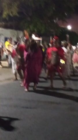 At night, a group of people are participating in a procession.  Most are barefoot. Three are dressed as Roman soldiers with red capes and helmets. One person is dressed as Jesus, wearing a tan undergarment and a maroon cape. The group is walking on a paved road, and several other people are gathered on the sidewalks. A few trees are visible in the background, as well as some buildings with bright lights. A motorcycle drives by at the beginning of the video.
