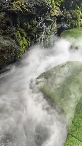 The media file showcases a lush, naturalistic setting with a small waterfall cascading down moss-covered rocks. A thick white mist rises from a dark pool of water, creating a dramatic and ethereal atmosphere. The mist obscures much of the water's surface, making it difficult to discern any specific objects within it. However, the mist also curls around two distinct, raised platforms covered in vibrant green artificial turf. These platforms are strategically placed within the scene, adding an element of curated design to the otherwise natural environment.

Ferns and other greenery hang from the rocks above, suggesting a tropical or humid climate, consistent with the reported location in Ban Na Chom Thian, Thailand. The lighting appears diffused, implying it might be a cloudy day or that the area is heavily shaded by the surrounding foliage. There are no people or animals visible in the frame. The overall mood conveyed is one of tranquility and perhaps a touch of mystery due to the pervasive mist. The mist's movement suggests a gentle breeze or the continuous flow of water. There is no visible text within the media file. The scene appears to be an outdoor installation, possibly a garden feature or a themed exhibit, designed to evoke a sense of natural wonder.