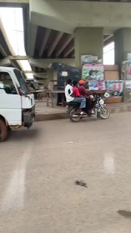 A white truck with a flat tire and a spare tire on top drives past a busy street market under an overpass. People are seen on motorcycles and at market stalls, with some activity around POS stands. The road surface is paved and appears somewhat worn. The environment is outdoors and appears to be a commercial area.