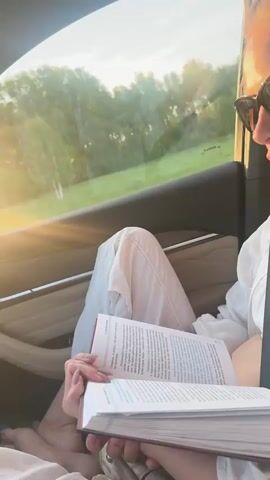 A woman is seen sitting in the passenger seat of a car, engaged in reading a book. She is wearing sunglasses, a white collared shirt, and light-colored pants, with a seatbelt fastened across her. Her hair is pulled back. Both hands are holding the open book, and her lips occasionally move as if she is reading aloud or silently mouthing the words. The text in the book appears to be in Russian.

Through the car window, a bright, sun-drenched landscape is visible, featuring green trees and fields, suggesting a rural or semi-rural setting. A strong sun flare emanates from the low sun, indicating either late afternoon or early evening (golden hour). The car's interior has light-colored upholstery and dark accents.

The woman's demeanor is calm and focused on her reading. She is heard speaking in Russian, saying, "ще потякивания. Нужно лечь на спину на ровную мягкую поверхность без подушки" (approximately: "more stretches. You need to lie on your back on a flat soft surface without a pillow"), suggesting the book contains instructions or information, possibly related to physical activity or health. The overall scene depicts a peaceful moment of reading while traveling.