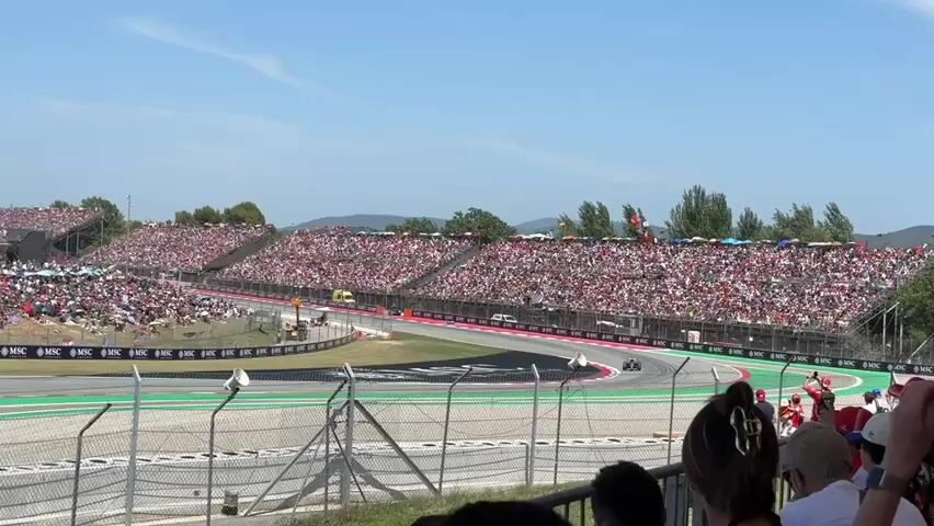 A race car is speeding around a track with a large crowd of spectators in the background. The spectators are seated in tiered grandstands that surround the track. The grandstands are filled with people wearing red and white clothing. The track is a paved surface with green and white markings. There are trees and hills in the background, and the sky is clear and blue. The race car is dark green and appears to be a Formula 1 car. The car is turning to the right as it moves around the track. There are several fences and barriers separating the spectators from the track. There are also some advertising banners visible along the side of the track. The video is shot from a low angle, behind a fence.