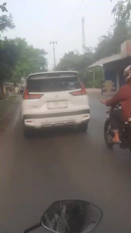 The video, filmed from the perspective of a person riding a motorcycle in Cae, Indonesia, shows a road scene during the daytime, possibly under hazy or overcast conditions. In the foreground, the handle and rearview mirror of the camera-holder's motorcycle are visible, with a reflection of what appears to be the rider's face or helmet in the mirror.

Ahead, a white Mitsubishi Xpander car with the license plate "B 1960 YCN" is being followed. To the right of the white car, another person in a reddish-brown jacket and helmet is riding a motorcycle, which bears the license plate "D 3584 MY". In the background, a yellow truck is partially visible, indicating other vehicles on the road. The sides of the road are lined with green trees and some small buildings or structures, with power lines and utility poles in the distance. The overall scene depicts a typical busy street in a semi-urban or rural area of Indonesia.